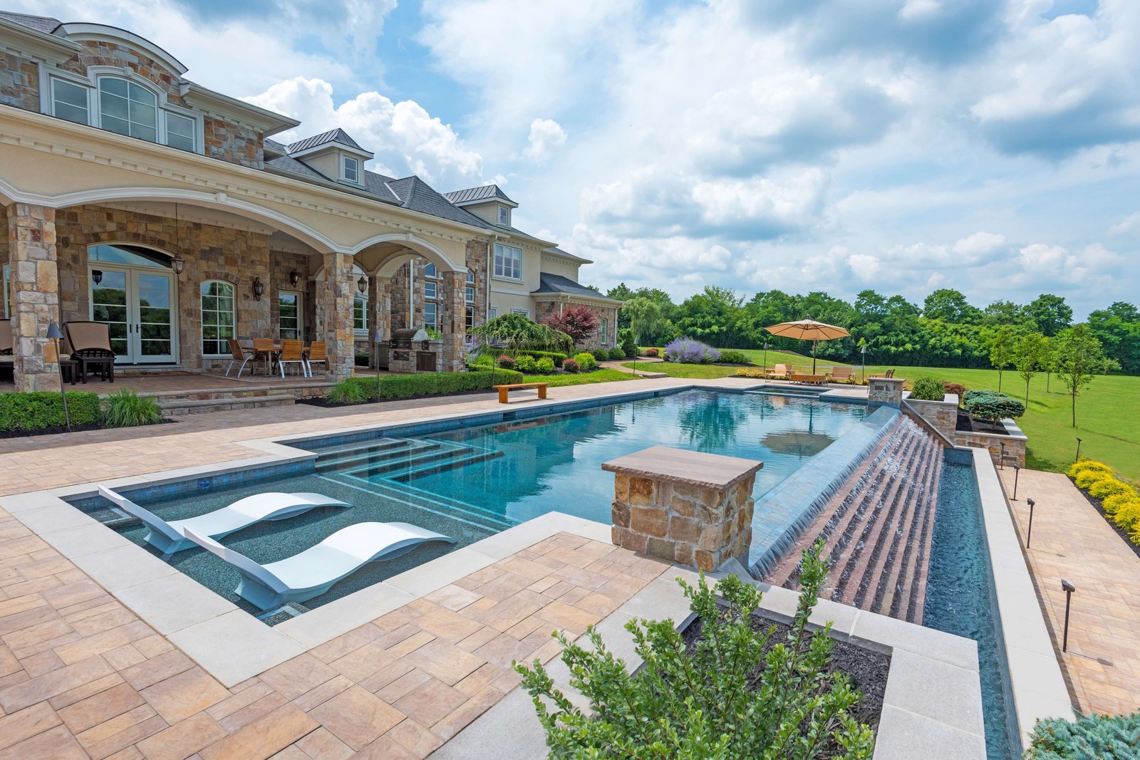 Seasonal World luxury inground pool installation in New Jersey &mdash; geometric pool with raised spa, tanning ledge, paver patio, and stone mansion behind